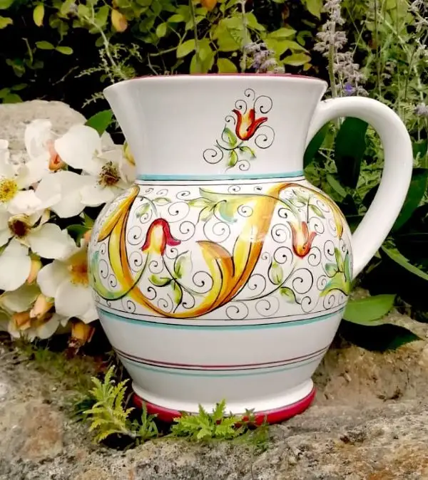 A pitcher from the Primizia dinnerware collection sitting on a rock surrounded by flowers. It's hand-painted in Deruta, Italy 