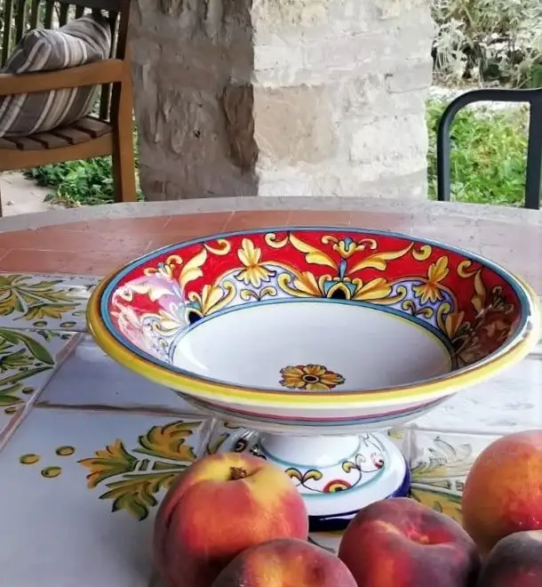 A footed ceramic fruit bowl hand-painted in an intricate red design is sitting on a table surrounded by peaches